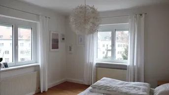 Helles Schlafzimmer mit zwei Fenstern, Holzboden und moderner Deckenleuchte, die viel Licht spendet.