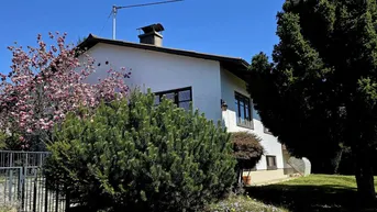 Weißes Einfamilienhaus mit gepflegtem Vorgarten und blühendem Baum unter blauem Himmel.