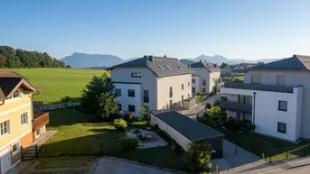 Luftaufnahme von modernen Wohnhäusern mit grünen Feldern und Bergen im Hintergrund unter blauem Himmel.