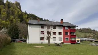 Mehrfamilienhaus mit roter und weißer Fassade, umgeben von Grünflächen und Hügeln.