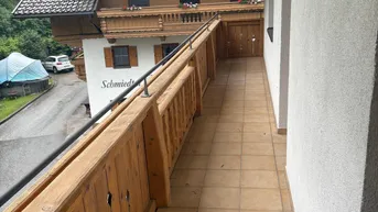 Langer Balkon mit Holzbalkonbrüstung und Blick auf ein traditionelles Gebäude und grüne Umgebung.
