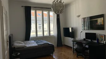 Helles Schlafzimmer mit Parkettboden, großem Fenster und einem funktionalen Arbeitsbereich.