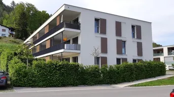 Modernes Mehrfamilienhaus mit weißer Fassade, Balkonen und gepflegter Hecke im Vordergrund.