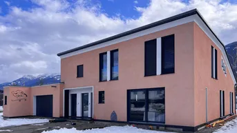 Modernes Einfamilienhaus mit rosafarbener Fassade und dunklen Fensterläden vor verschneiter Bergkulisse.