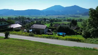 Weitläufige grüne Landschaft mit Häusern, einer Straße und Bergen im Hintergrund unter blauem Himmel.