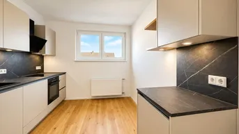 Moderne Einbauküche mit hellen Schränken, dunkler Arbeitsplatte und Holzfußboden, gut beleuchtet.
