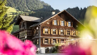 Traditionelles Holzhaus in den Bergen mit grüner Umgebung und blühenden Pflanzen im Vordergrund.