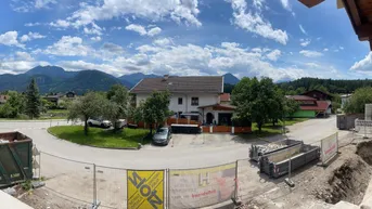 Panoramablick auf eine Baustelle mit Containern und Absperrungen vor einer Bergkulisse.