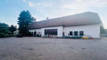 Außenansicht eines Gewerbegebäudes mit großem gepflasterten Vorplatz unter bewölktem Himmel und Bäumen.
