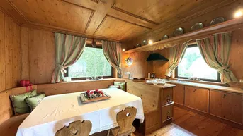 Traditionelle Holzküche mit Essbereich, Holztäfelung und Blick ins Grüne durch die Fenster.