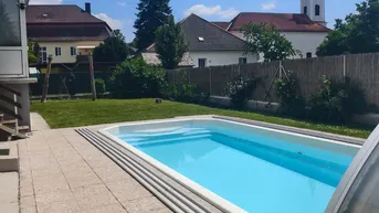 Außenpool mit Überdachung, umgeben von einer gepflasterten Terrasse und Rasenfläche mit Kirchenblick.
