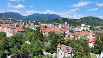 Panoramablick über die Stadt mit grünen Hügeln und Bergen im Hintergrund unter blauem Himmel.