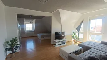 Helles und geräumiges Wohnzimmer mit Dachschrägen, Holzboden und einem angrenzenden Schlafbereich.