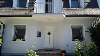 Weiße Hausfassade mit Mansarddach, kleinen Balkonen und gepflegtem Vorgarten unter blauem Himmel.