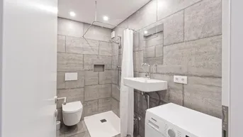 Modernes Badezimmer mit grauen Fliesen, Dusche, WC und integrierter Waschmaschine.