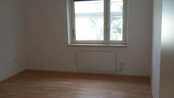 Leeres Zimmer mit hellem Laminatboden, weißen Wänden, Fenster und Heizkörper.