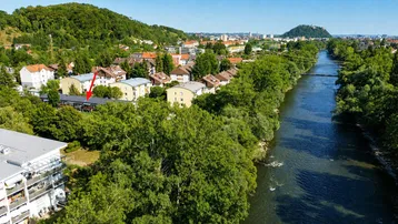 Luftaufnahme eines Wohngebiets entlang eines Flusses mit viel Grün und Stadtsilhouette im Hintergrund.