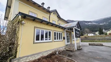 Gelbes Gebäude mit weißen Fenstern und einem Vordach, umgeben von einer ländlichen Landschaft mit Bergen im Hintergrund.
