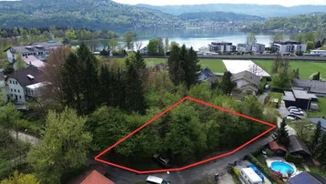 Luftaufnahme eines Baugrundstücks mit roter Markierung und Blick auf den See und die umliegende Landschaft.