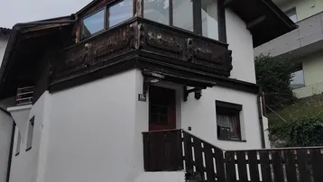 Traditionelles Gebäude mit weißer Fassade, Holzbalkonen und einem spitzen Dach vor bergiger Kulisse.