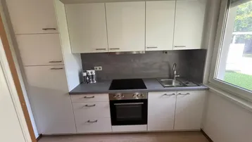 Moderne Küche mit weißen Hochglanzschränken, grauer Arbeitsplatte und Blick ins Grüne.