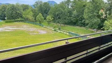 Weitläufiger Blick vom Balkon über grüne Wiesen, einen kleinen Bach und bewaldete Berge im Hintergrund.
