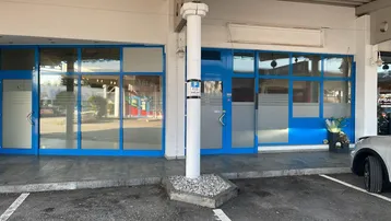Vorderansicht eines Geschäftsgebäudes mit großen blauen Fensterfronten und Eingangstüren sowie einem Parkplatz davor.
