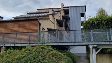 Außenansicht des Gebäudes mit erhöhtem Zugangssteg und üppiger Bepflanzung im Vordergrund.