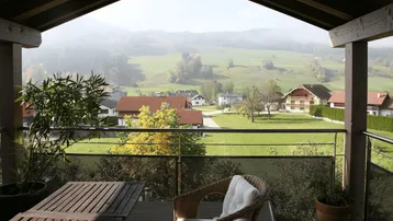 Überdachter Balkon mit Holztisch, Korbstuhl und weitem Blick über die grüne Landschaft.