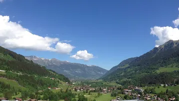Panoramablick auf ein grünes Tal mit Bergen und einem Dorf unter blauem Himmel.