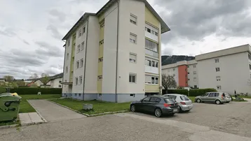 Mehrfamilienhaus mit hellgelber Fassade, Parkplätzen und Grünflächen unter bewölktem Himmel.