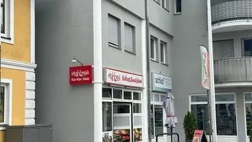Graues Mehrfamilienhaus mit Geschäftsräumen im Erdgeschoss und Wohnungen darüber, an einer Straße gelegen.