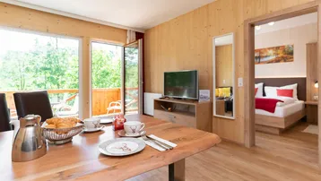 Gemütlicher Wohn-Essbereich mit Holzwänden, Esstisch und Zugang zu einem Balkon mit grüner Aussicht.