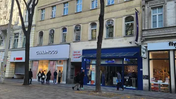 Gebäude an einer belebten Straße mit Geschäften im Erdgeschoss und Wohnungen darüber, umgeben von Bäumen.