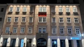Nachtaufnahme eines Mehrfamilienhauses mit beleuchteter Fassade und Geschäftsflächen im Erdgeschoss.