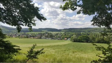 Weitläufige Landschaft mit einem Dorf, grünen Feldern und Wäldern unter bewölktem Himmel.