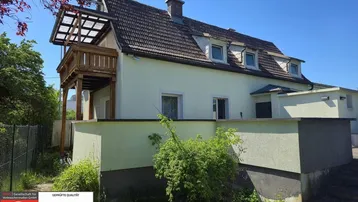 Ansicht des Hauses mit einem erhöhten Holzbalkon und einer hellen Fassade unter blauem Himmel.