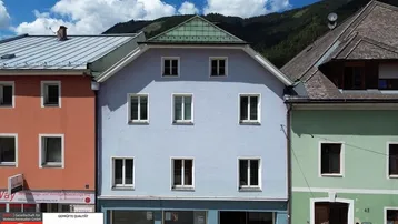 Vorderansicht eines hellblauen Gebäudes mit Geschäftsflächen im Erdgeschoss, umgeben von anderen farbigen Häusern und Bergen.