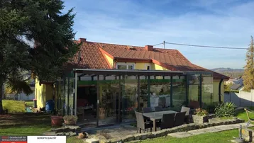 Hausfassade mit verglastem Wintergarten, davor eine möblierte Terrasse und weitläufiger Gartenbereich.