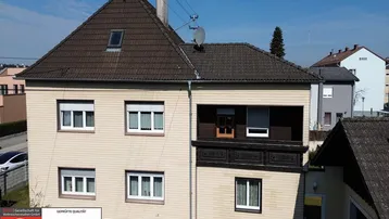 Vorderansicht eines Mehrfamilienhauses mit mehreren Fenstern und einem kleinen Balkon unter blauem Himmel.
