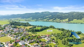 Luftaufnahme eines Dorfes am Seeufer mit umliegenden Hügeln und Bergen.