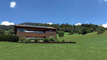 Haus in ländlicher Umgebung mit grünen Wiesen und bewaldeten Hügeln unter blauem Himmel.