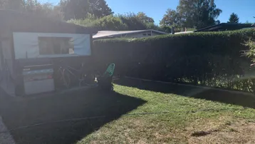 Sonnige Gartenfläche mit Rasen, einer hohen Hecke und einem Grillbereich neben einem Schuppen.