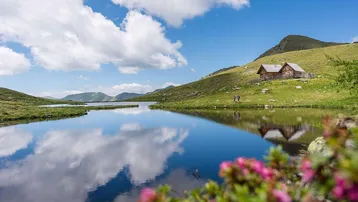 Malerische Berglandschaft mit klarem See, grünen Hügeln und traditionellen Holzhütten unter blauem Himmel.