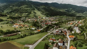 Luftaufnahme eines malerischen Dorfes in einem grünen Tal, umgeben von bewaldeten Bergen und einer Straße.