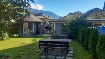 Sonniger Garten mit Sitzbereich, Grillplatz und einem Pizzaofen im Freien vor Bergkulisse.