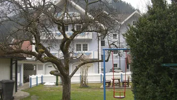 Großer Gartenbereich mit Schaukel, altem Baum und Blick auf das Mehrfamilienhaus und die Berge.
