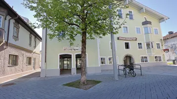 Historisches Gebäude mit hellgrüner Fassade und einem Baum davor, zeigt einen Eingangsbereich für ein Geschäft.