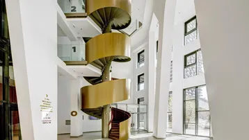 Große, helle Eingangshalle mit einer beeindruckenden goldenen Wendeltreppe und hohen Decken.