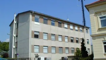 Mehrstöckiges Gebäude mit beiger Fassade und vielen Fenstern an einer Straße. Ein Mobilfunkmast ist im Hintergrund sichtbar.
